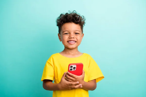 ターコイズカラーの背景に隔離されたスマートフォンを保持するスタイリッシュなTシャツを着用した満足のいく小さな男の子の肖像 — ストック写真