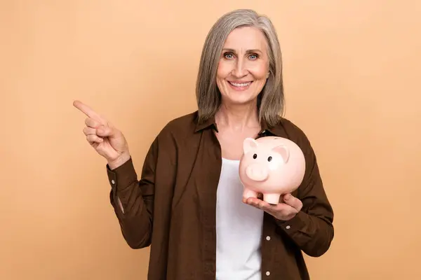Photo of cheerful grandmother hold money bank pig indicate finger empty space isolated on beige color background.
