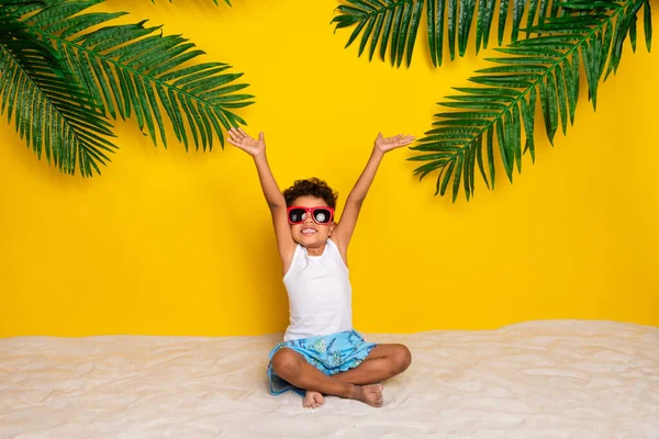 全长照片 迷人无忧无虑的小男孩身穿白衬衫 深色眼镜 坐着沙子 举起双手 臂膀孤立的黄色背景 — 图库照片