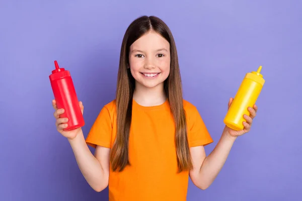 照片上的笑容可亲的女生穿着橙色T恤烹饪用番茄酱与芥末分离的紫色背景 — 图库照片