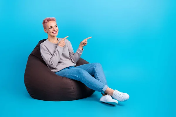 Photo of cheerful nice adorable woman look directing at offer empty space sit on comfortable pouf isolated on blue color background.