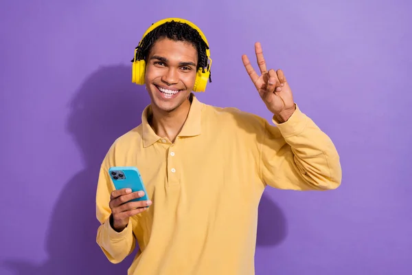 Photo of cheerful funny person with cornrows listen music in headphones hold smartphone show v-sign isolated on purple color background.