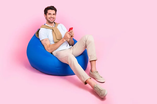 ピンク色の背景に隔離された快適なビーンバッグテキストスマートフォンユーザーのリラクゼーションに横たわる陽気な男の全身の長さ — ストック写真