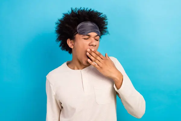 Portrait of young exhausted sleepy person yawning arm cover mouth blindfold mask forehead isolated on blue color background.