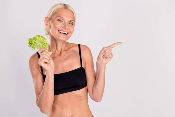 Portrait of gorgeous aged lady hold fresh lettuce direct finger empty space think waist self care isolated on grey color background.