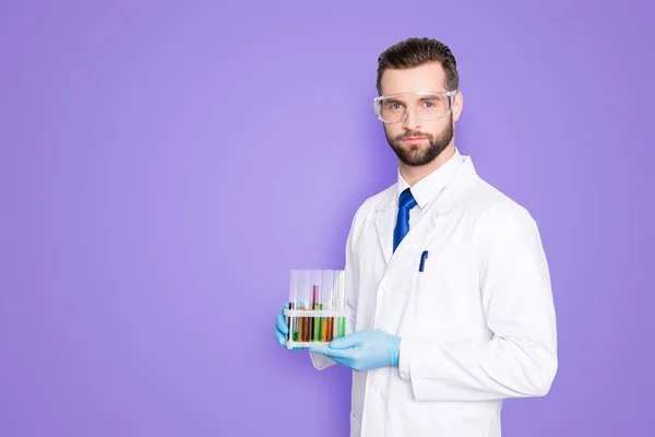 Portrait with copy space, empty place of attractive stylish scientist with bristle in white lab coat, tie holding test tubes with multi-colored liquid, looking at camera over grey background.