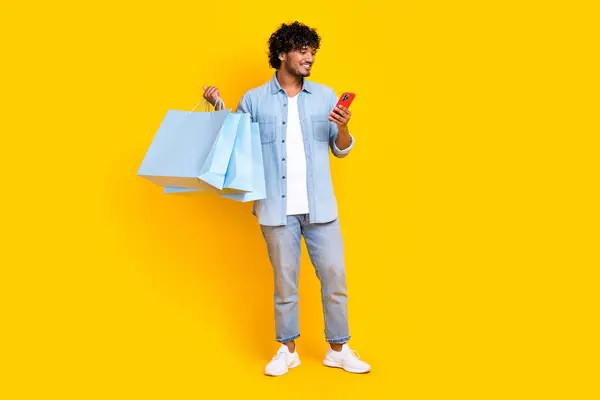 Full length photo of satisfied man with wavy hair wear jeans shirt hold new clothes look at smartphone isolated on yellow color background.