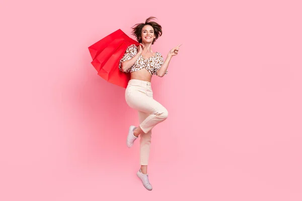 Full length photo of attractive woman wear top pants jumping hold shopping bags indicating empty space isolated on pink color background.