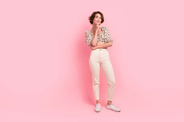 Full length photo of optimistic minded woman wear print top white pants look empty space hand on chin isolated on pink color background.