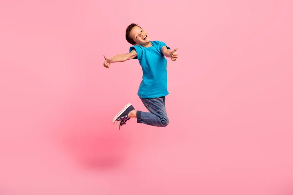 Full length photo of excited cool little boy dressed blue t-shirt jumping high showing two thumbs up isolated pink color background.