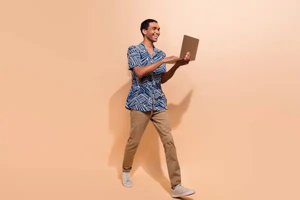 聪明而快乐的男人穿着印花衬衫的全副照片棕色裤子看笔记本电脑空旷的地方孤立在米色的背景下 — 图库照片