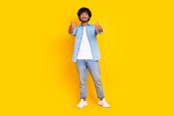 Full length photo of satisfied man with wavy hairstyle wear jeans shirt showing thumbs up good job isolated on yellow color background.