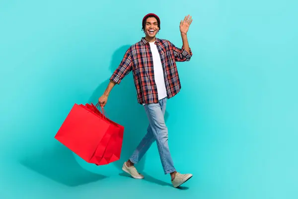 Full size portrait of cheerful cool person hold mall shop bags walking arm palm waving isolated on turquoise color background.