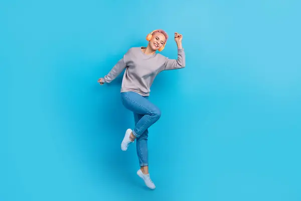 Full length photo of overjoyed lovely person jumping have good mood listen music headphones isolated on blue color background.