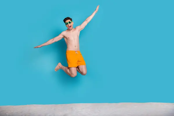 Full size photo of attractive young man jump flying arms plane wear trendy yellow shorts isolated on blue color background summer vacation.