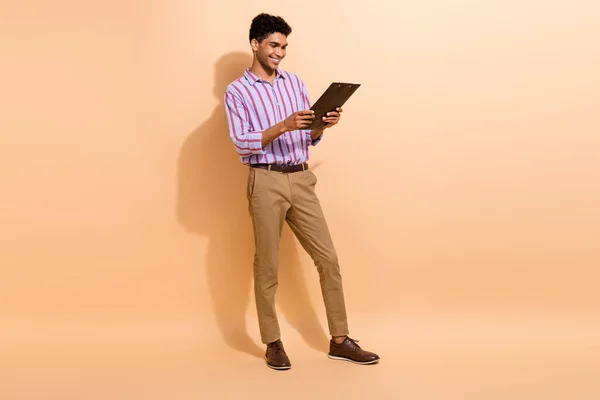 楽観的なクールな男のフルサイズの写真は ストライプのシャツブラウンパンツを見て ベージュ色の背景で隔離されたレポートを読みます — ストック写真