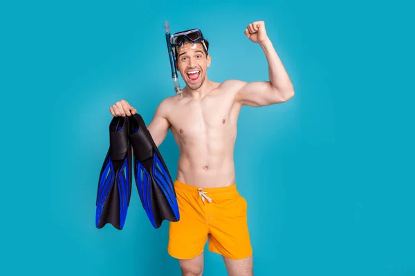 Photo of excited lucky shirtless man smiling rising fist holding flippers diving goggles tube isolated blue color background.