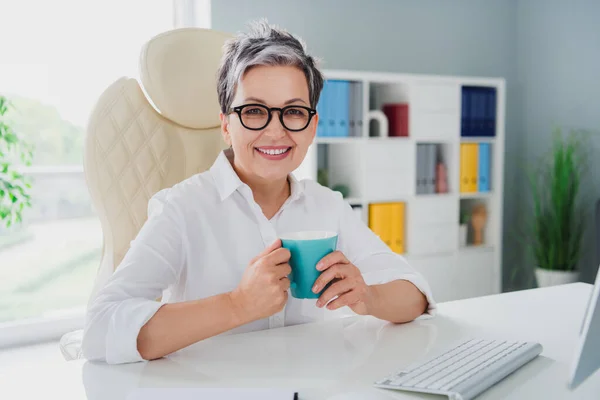 照片上漂亮而满意的成熟女人穿着白色的正式衬衫 在办公室窗口的浅色背景上与等客人隔离喝咖啡 — 图库照片