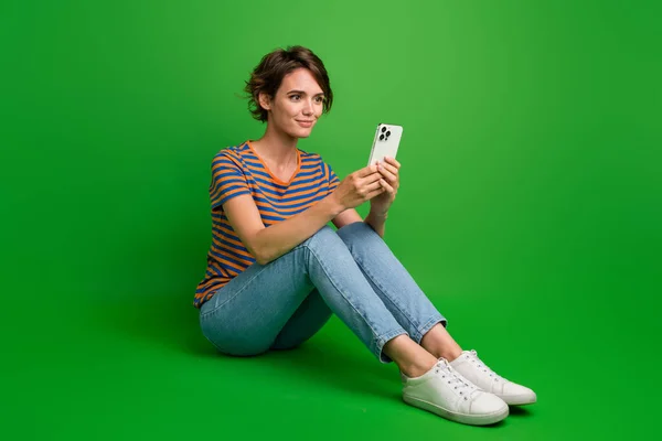 Full body photo of pretty positive lady sit floor use smart phone typing blog post isolated on green color background.