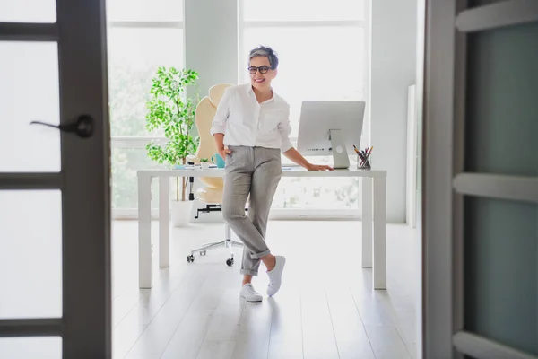Full body cadre of satisfied business lady formal shirt gray pants confident posing model isolated on workstation window light background.