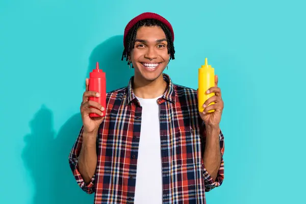Photo of toothy beaming nice guy with cornrows dressed checkered shirt hands hold mustard ketchup isolated on teal color background.