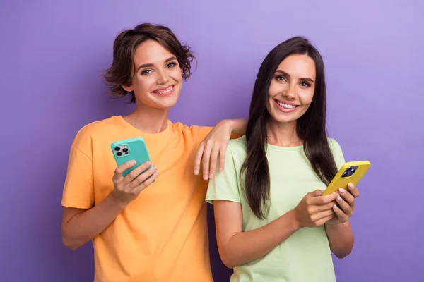 Photo of funky cute girls dressed t-shirts online typing sms apple samsung iphone devices isolated purple color background.