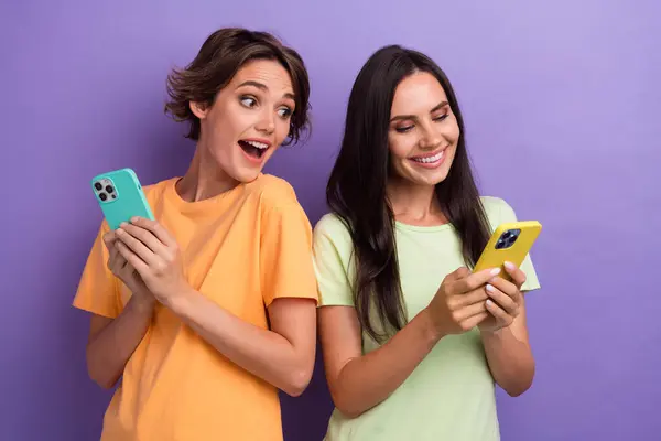 Photo of impressed funky girls dressed t-shirts online shopping modern devices isolated purple color background.
