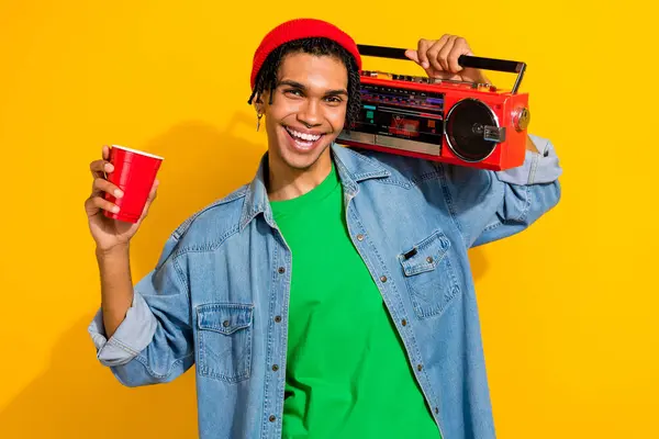 Photo of cheerful glad man music lover wear jeans shirt earrings listen audio hold plastic cup isolated on vivid yellow color background.
