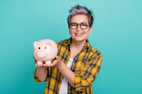 Photo of charming grandmother hold pig bank collect money dressed stylish checkered yellow outfit isolated on aquamarine color background.