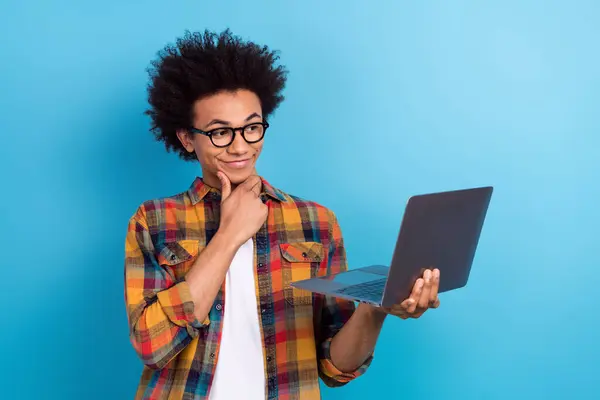 Portrait of minded pensive guy with afro hairstyle touch glasses look at laptop hand on chin read email isolated on blue color background.