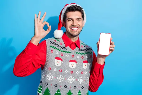 面白い男の写真は 青い色の背景に隔離されたスマートフォンディスプレイ上のアプリを推奨クリスマスの帽子を着用します — ストック写真