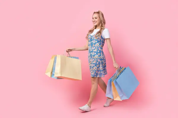 Full size profile photo of lovely positive girl hold shop bags walk empty space isolated on pink color background.