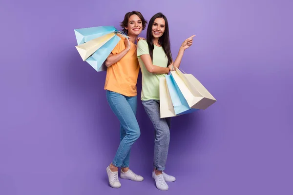 Full body photo brand logo advert youngsters girlfriends pointing finger mockup with bargain bags isolated on purple color background.