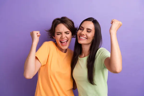 Photo of lucky excited ladies wear t-shirts hugging yelling yes rising fists isolated violet color background.
