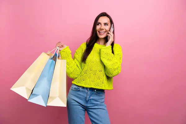 Portrait of good mood woman wear yellow pullover talk on smartphone hold shopping bags look empty space isolated on pink color background.