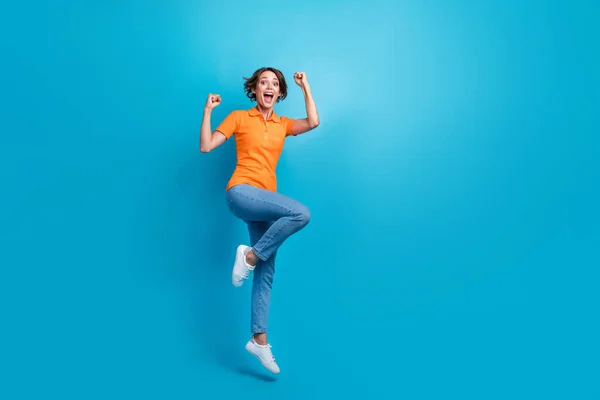 満足した女性のフルサイズの写真は オレンジ色のTシャツスタイリッシュなジーンズを着用します ジャンプショット ブルーカラーの背景に隔離されたベット — ストック写真