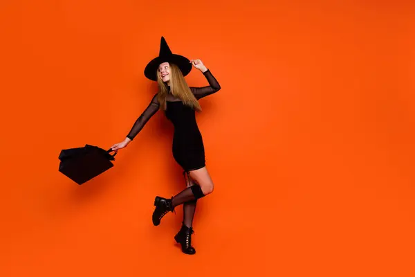 スイートケアフリーの女性の全長の写真は 買い物客を保持する黒いウィザードの衣装の帽子を着用します 空のスペース 隔離されたオレンジ色の背景 — ストック写真