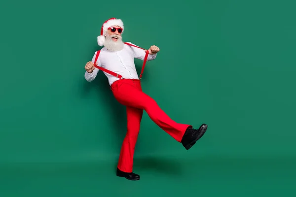 Full length photo of cool man dressed xmas costume eyewear adjusting suspenders walking empty space isolated green color background.