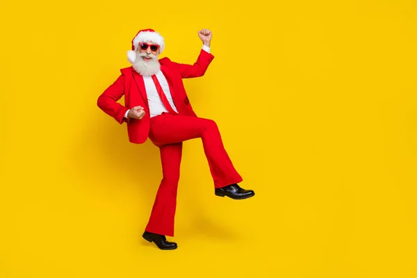 Full length photo of attractive lucky retired guy wear red tux claus hat walking rising fists empty space isolated yellow color background.