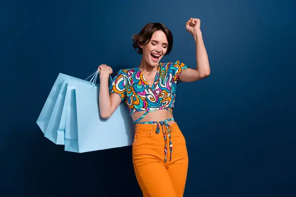 Photo of attractive young woman raise fist celebrate shopping wear trendy flared pants clothes isolated on dark blue color background.