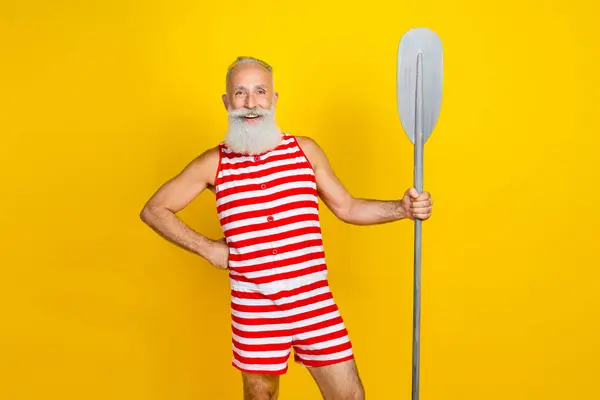 Photo of funny bearded old sportsman wear striped swimsuit holding boat oar when swimming in river isolated on yellow color background.