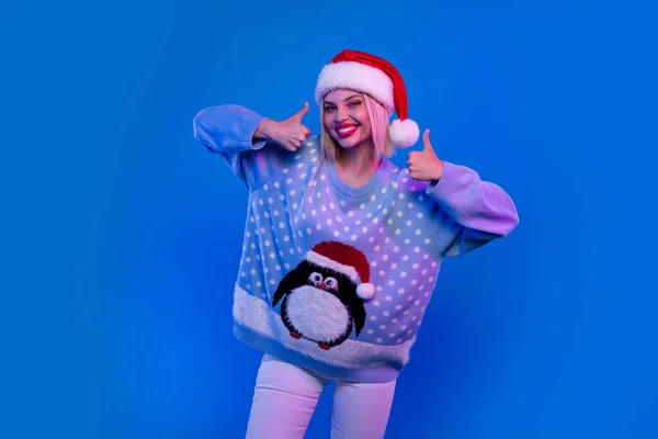 Photo of adorable funky lady santa helper dressed ugly print xmas sweater showing two thumbs up isolated blue color background.