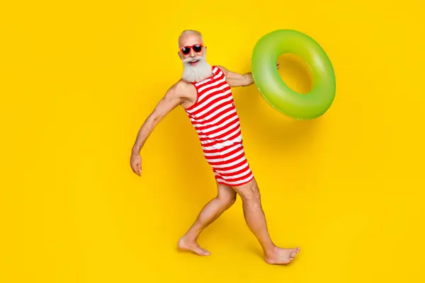 Full length photo of funky good mood elderly guy dressed striped red swimsuit walking swim buoy isolated yellow color background.