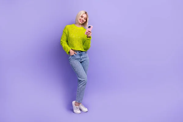全长照片甜美闪亮的女人穿着针织毛衣发短信现代装置空旷的空间孤立的紫罗兰色背景 — 图库照片