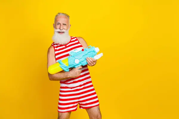 Photo of brutal crazy grandfather red striped swim shorts overalls splashing water gun at swimming pool isolated on yellow color background.