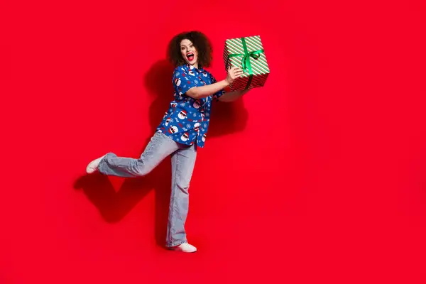 漂亮的年轻女子拿着礼品盒 穿着时尚的蓝色圣诞印花衣服 背景为红色 全长照片 — 图库照片