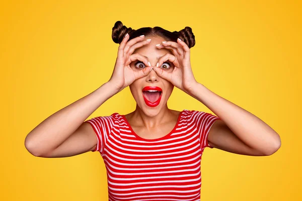 Pretty girl with surprised face holding fingers near eyes like glasses: mask like super hero or owl isolated on red background.