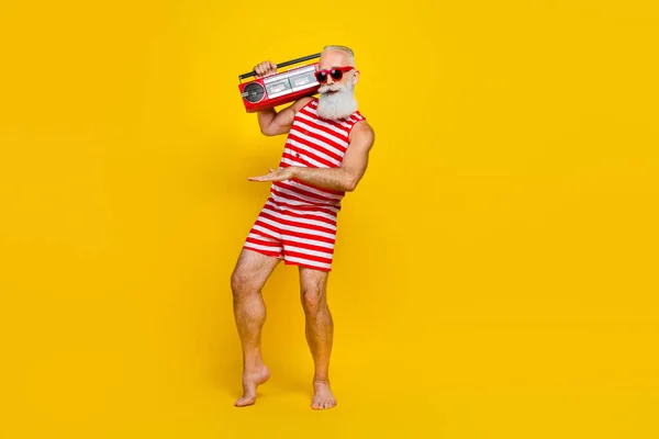 Full length photo of funny man in striped swimsuit dancing at the swimming pool dj hold boombox isolated on yellow color background.