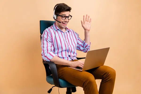 Photo of cheerful glad guy sitting office chair speaking saying hello earphones microphone video call isolated on beige color background.