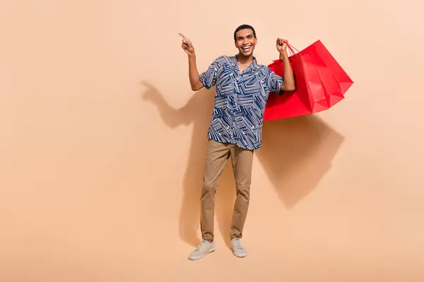 Full length photo of optimistic man dressed shirt trousers hold shopping bags directing empty space isolated on beige color background.